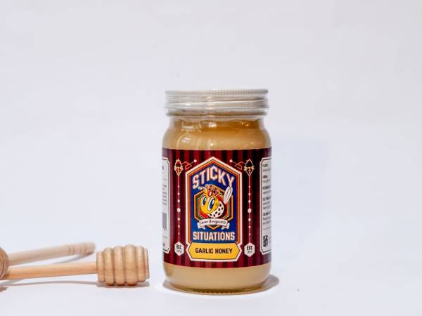 Image of 8 oz. sticky situations garlic honey with a honey dipper to the left.