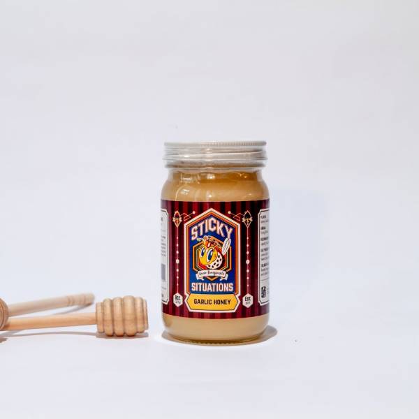 Image of 8 oz. sticky situations garlic honey with a honey dipper to the left.